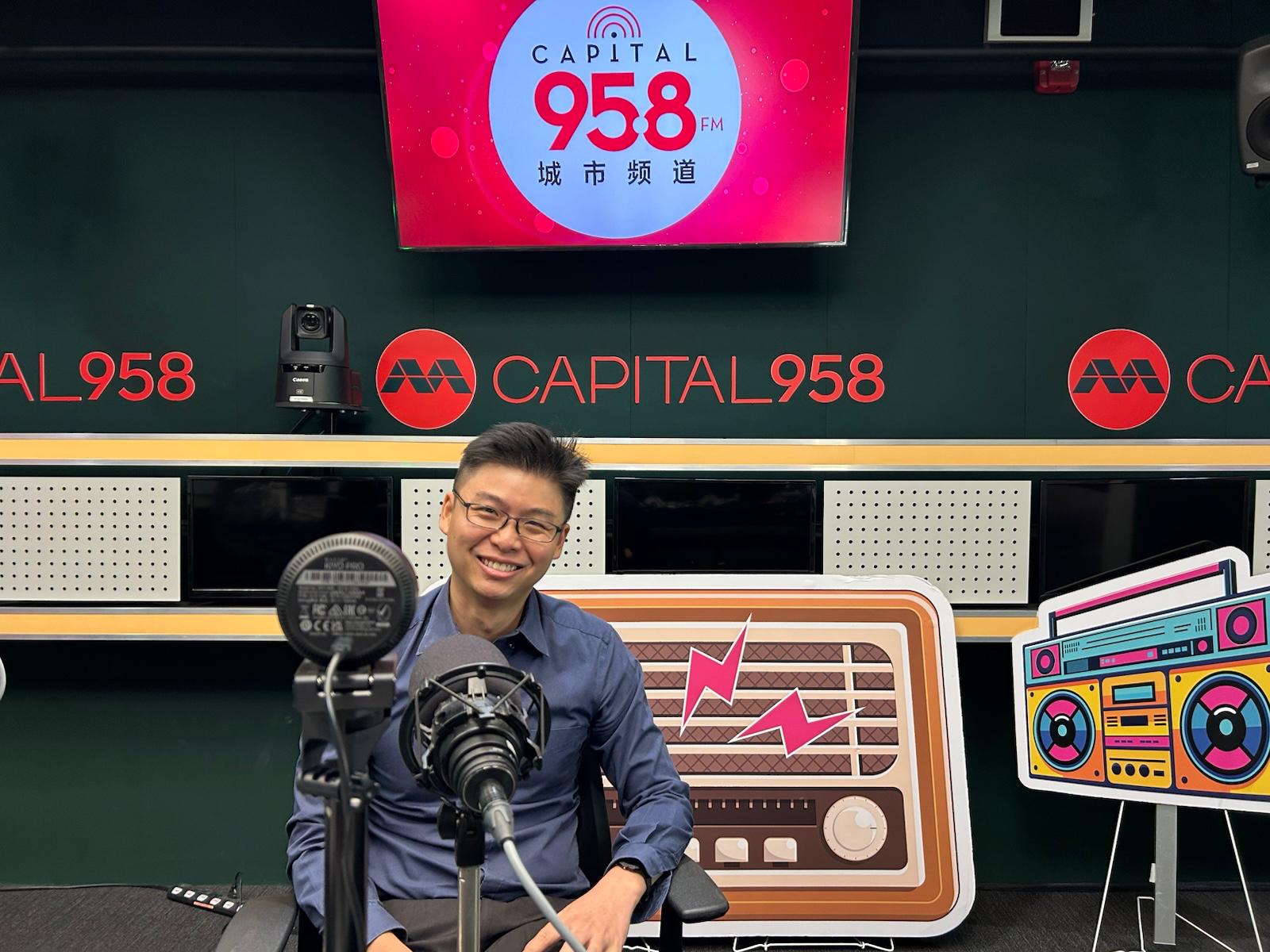 Smiling man in a blue shirt sits at a microphone in a Capital 958 radio studio with branding on the wall behind him.