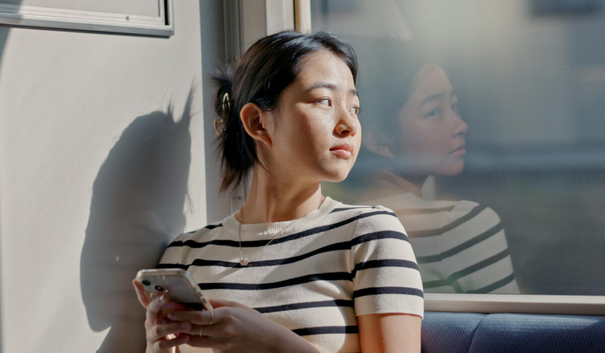 Woman in a striped shirt sits by a window, gazing outside while holding a smartphone.