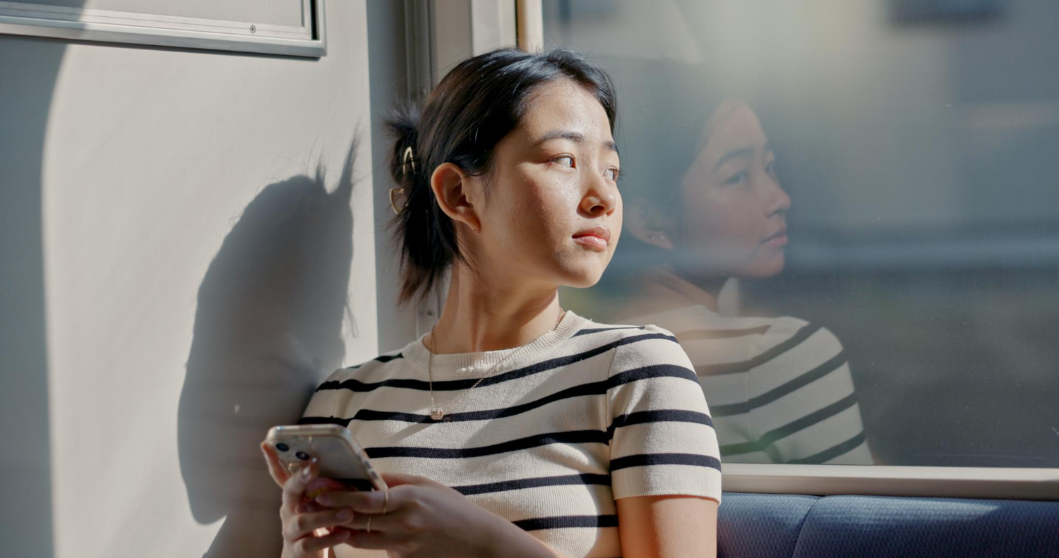 Woman in a striped shirt sits by a window, gazing outside while holding a smartphone.