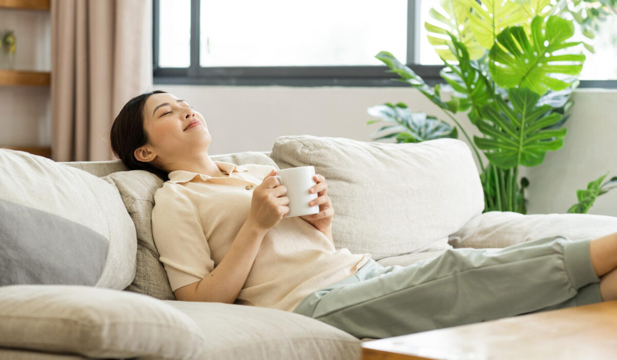 Woman relaxing on a beige sofa indoors, holding a white mug and smiling, sunlit window and green plants in the background.