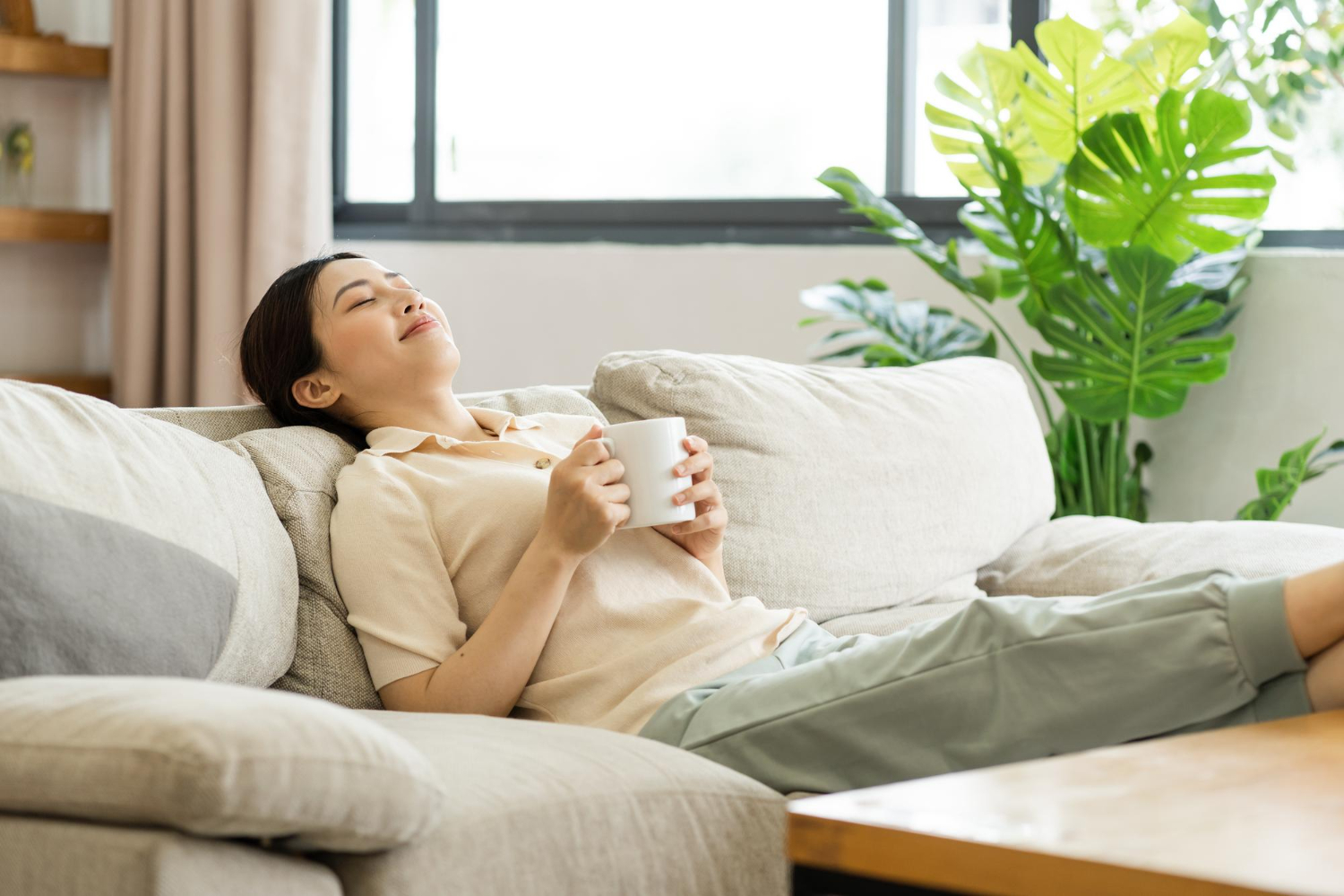 Woman relaxing on a beige sofa indoors, holding a white mug and smiling, sunlit window and green plants in the background.