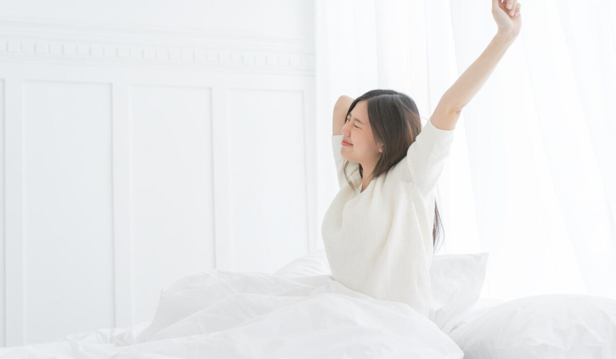 Woman in a white sweater sits up in bed and stretches with arms raised in a bright white bedroom, smiling.