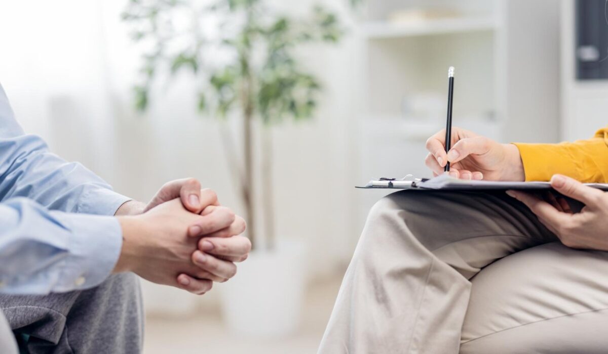 Therapist taking notes on a clipboard while client sits with clasped hands in a counseling session.