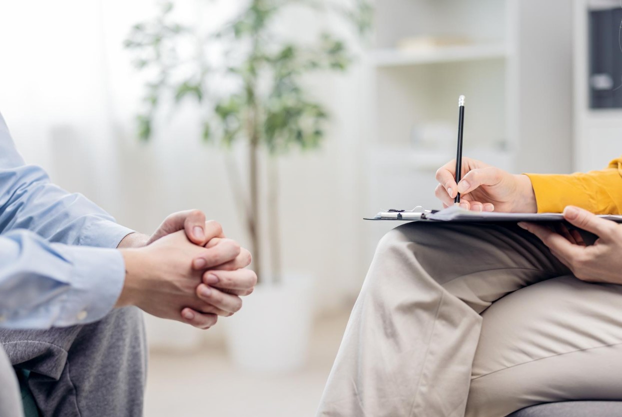 Therapist taking notes on a clipboard while client sits with clasped hands in a counseling session.
