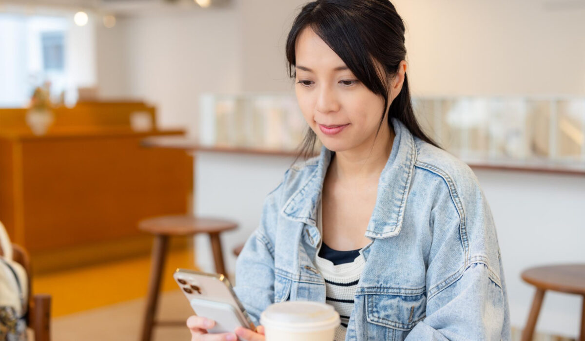 Woman in a denim jacket checks her phone at a cafe table with a takeaway coffee cup nearby.