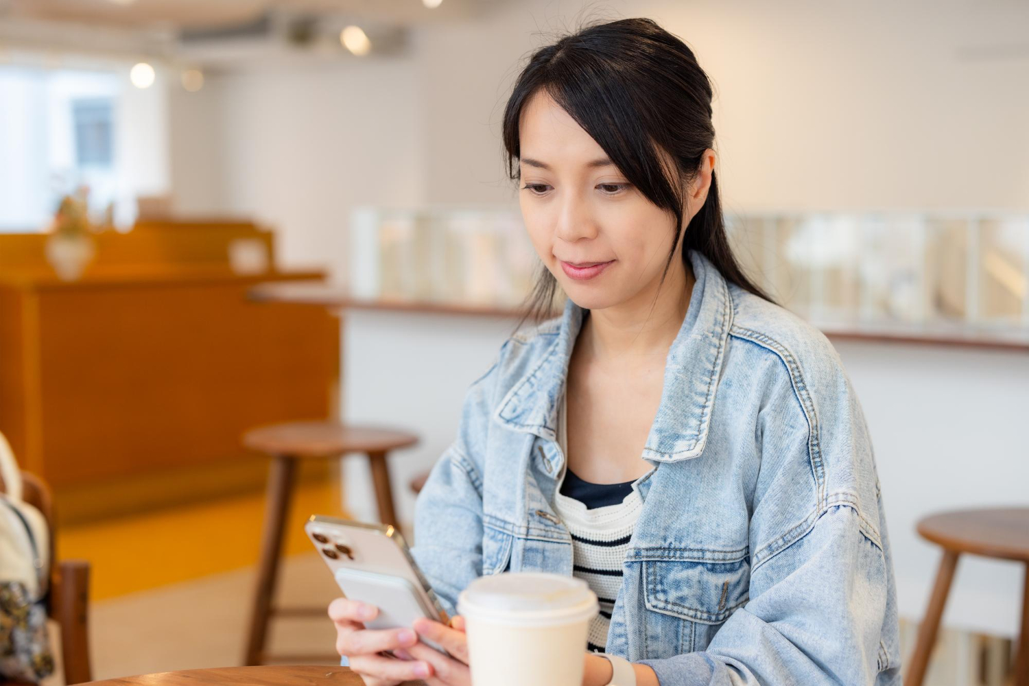 Woman in a denim jacket checks her phone at a cafe table with a takeaway coffee cup nearby.